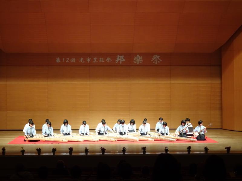 舞台上の第12回光市芸能祭 邦楽祭と書かれた看板の下で赤い絨毯の上で琴を演奏する16人の女子学生と三味線を演奏する2人の女子学生の写真