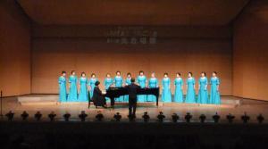 舞台上で黒い服を着て指揮をする男性を先頭にピアノを演奏する女性の奥に立っている水色のドレスを着た14人の女性の写真