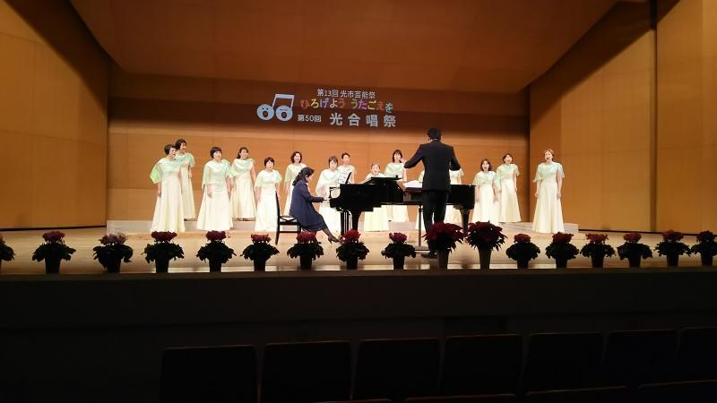 壁に「ひろげよううたごえを 第50回光合唱祭」と書かれた舞台の中央で合唱する15人の白い衣装を着た女性の前でピアノを演奏する女性と指揮をする男性の写真