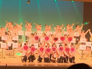 舞台の奥では吹奏楽団員が演奏をしていて、手前では吹奏楽団員が二列に並んでうちわを持っている写真