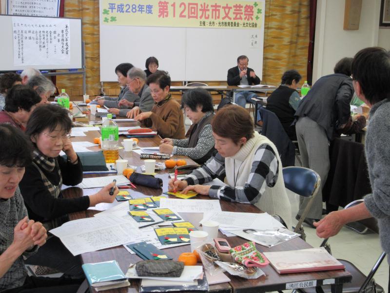 平成28年度第12回光市文芸祭と書かれたパネルの前の席に座っている男性とその前に並んでいる長机で紙を広げながら書き物をしている14人の男女の写真