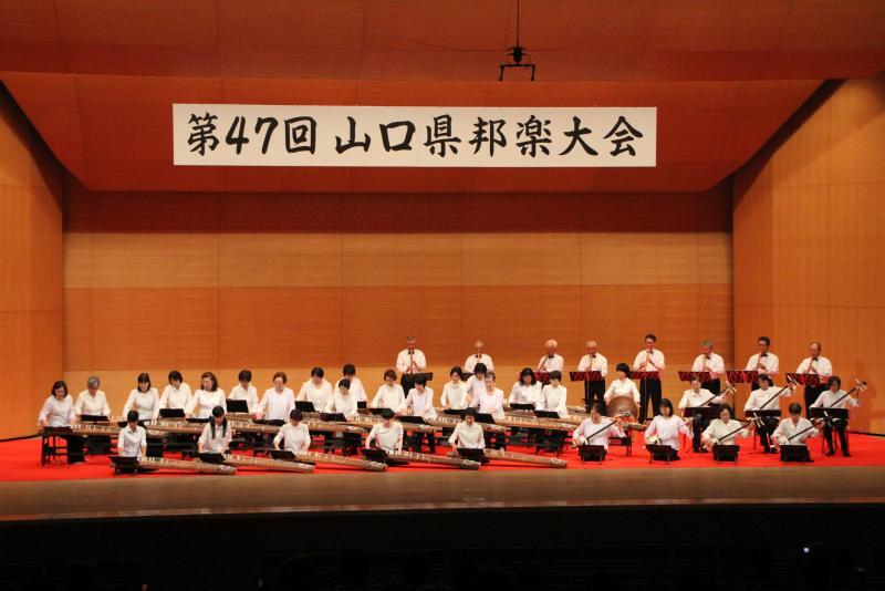 舞台の上で琴、尺八、三味線、太鼓を演奏している38人の男女の写真