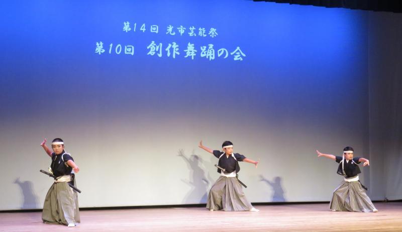 青いライトを背景に白い文字で第14回光市芸能祭 第10回創作舞踏の会と映し出された舞台のスクリーンの前で腰に剣を差して踊る黒い袴姿の3人の男の子の写真