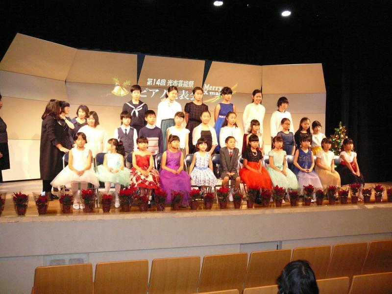 4人の女性が端に立っている舞台の真ん中で正装で3列に並んでいる27人の幼稚園から中学生くらいまでの年代の子供たちの写真