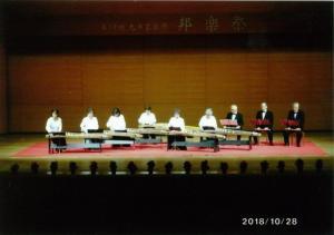 舞台上で琴を演奏する白い服の6人の女性と尺八を吹く黒い服の3人の男性の写真