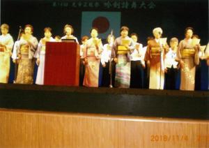 吟剣詩舞大会という文字と日本国旗が映し出されたスクリーンを背景に舞台上で黄色い紙を持って立っている着物姿の15人の男女の写真