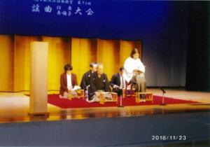 青いライトを背景に白い文字で謡曲大会と書かれたスクリーンの前に立っている金屏風の前の赤い絨毯の上に座っている5人の男女の写真