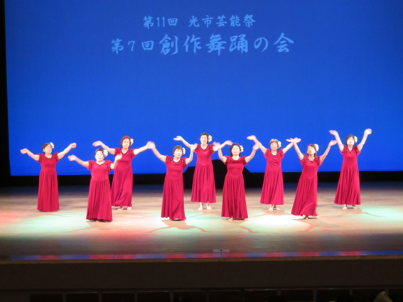 青いライトを背景に白い文字で第11回光市芸術祭 第7回創作舞踏の会と映し出されたスクリーンの前で両手を挙げている赤いドレスを着た9人の女性の写真