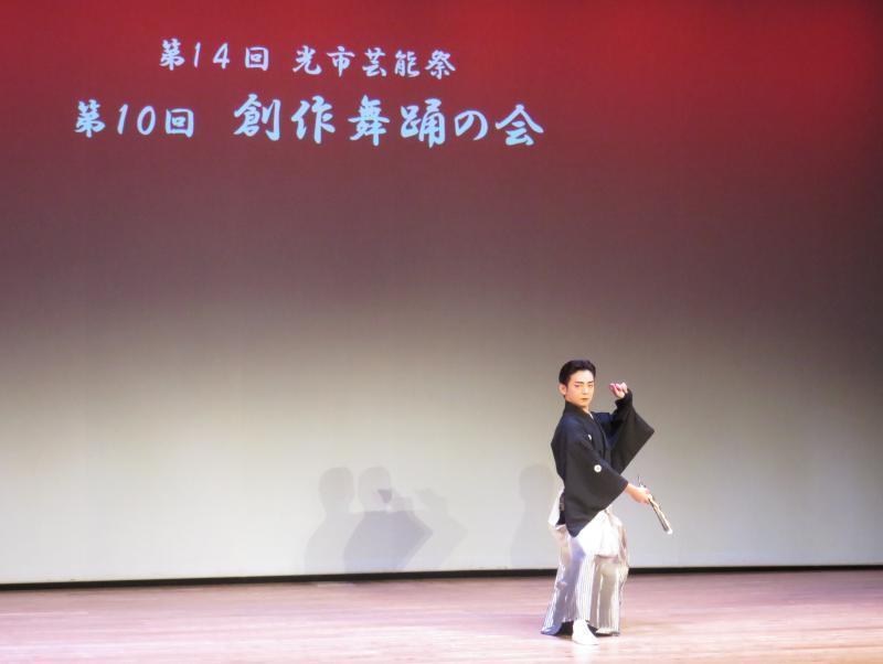 赤いライトを背景に白い文字で第14回光市芸能祭 第10回創作舞踏の会と映し出された舞台のスクリーンの前で扇子を持って踊る黒い袴姿の男性の写真
