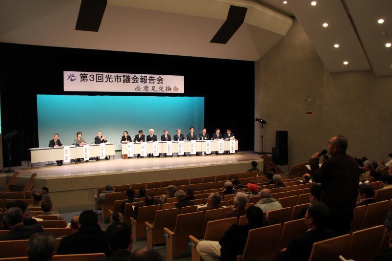 市民ホールで壇上にいる議員数名にマイクを使って質問する聴講者の写真