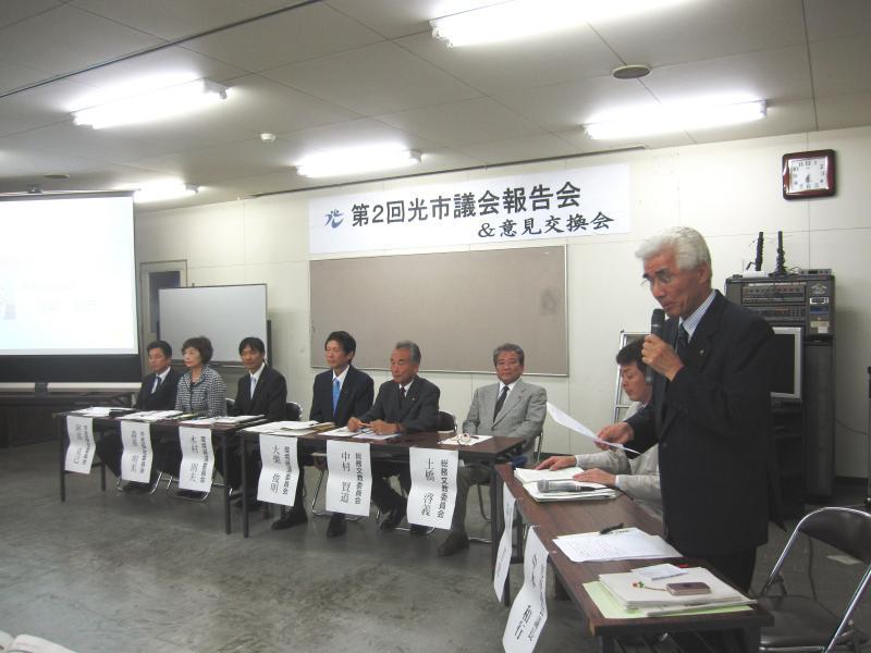 一列に並べた会議テーブルに8人の議員が座り報告会に臨んでいる写真