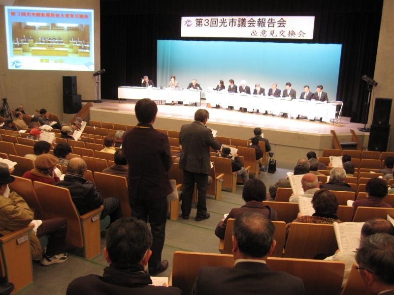 第3回光市議会報告会にて、壇上にいる議員数名に向け、立ち上がってマイクで意見を伝える参加者の写真