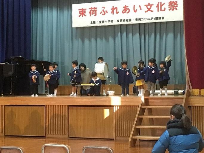 ステージで束荷幼稚園の園児たちがタンバリン、トライアングル、太鼓、鈴、シンバル、ピアニカで合奏しているのを舞台の下から先生が見守っている写真