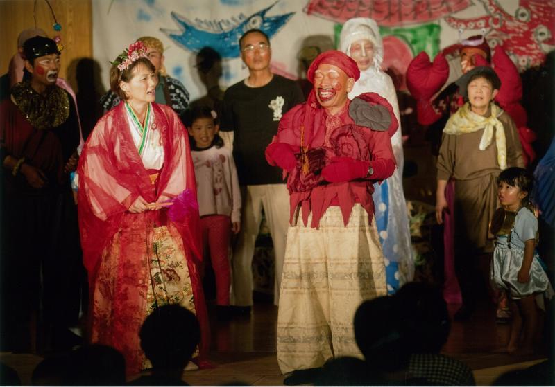 顔に化粧をして赤い帽子、手袋、上着を着た男性と、和服の衣装を着た女性が演劇を披露している写真