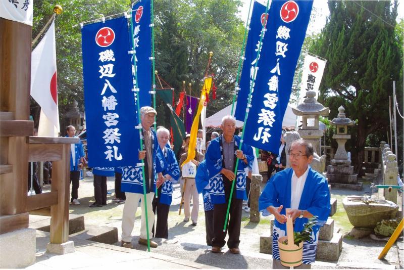 法被を着て磯辺八幡宮祭礼と書かれたのぼり旗を持つ男性と、榊の入った桶を持つ男性の写真