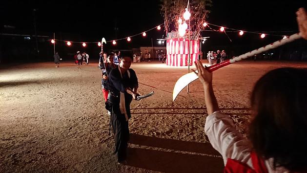 奥に盆踊りの櫓が立っている祭り会場で、衣装を着て刀を持って互いに向かい合っている男女の写真