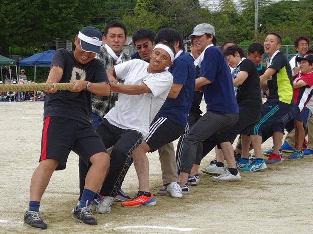 白いハチマキをした男性たちがグラウンドで歯を食いしばりながら綱引きをしている写真