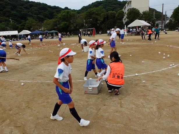 玉入れの後片付けをしている隊員の横を帽子に鉢巻きを付けた体操服姿の小学生たちが通っている写真