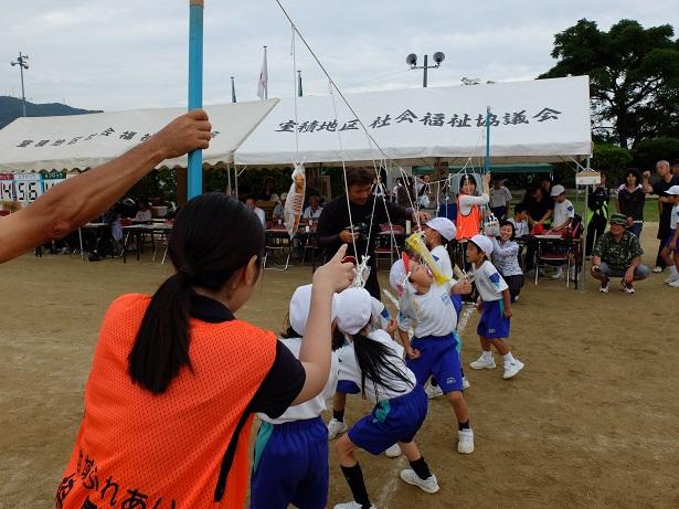 帽子に体操服姿の小学生たちがパン食い競争でパンを口にくわえようとしていて、そのパンがぶら下がっている棒を隊員が支えている写真