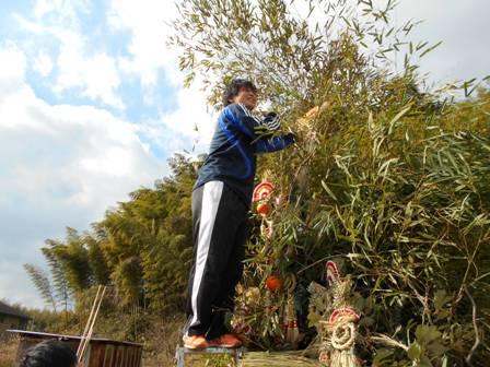 外に置いてある竹で組まれた大きなヤグラの前で男性が脚立に上ってしめ縄を取り付けている写真