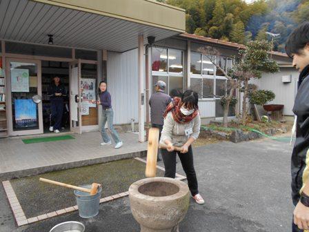 公民館の外で杵で餅をついている女の子を見守っている男性スタッフの写真