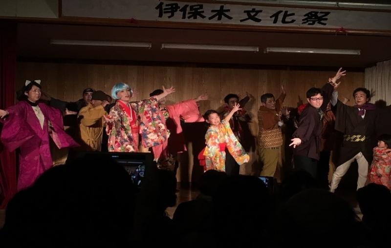 舞台上で和服の衣装を着て両手を広げてポーズをとる出演者たちの集合写真