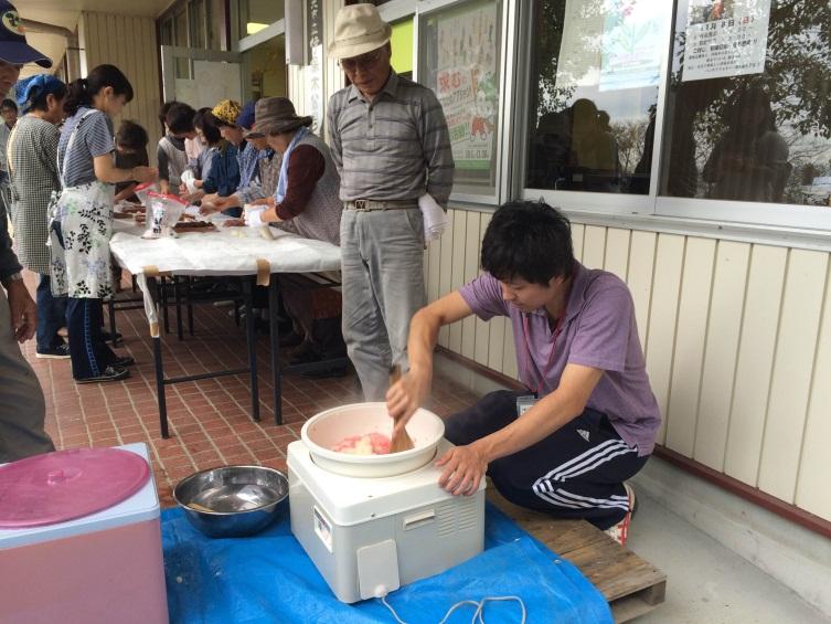 公民館で餅つき機の中味をしゃもじで混ぜている男性を帽子を被った男性が見ている。その横のテーブルでエプロン姿の女性たちが餅を丸めてる写真