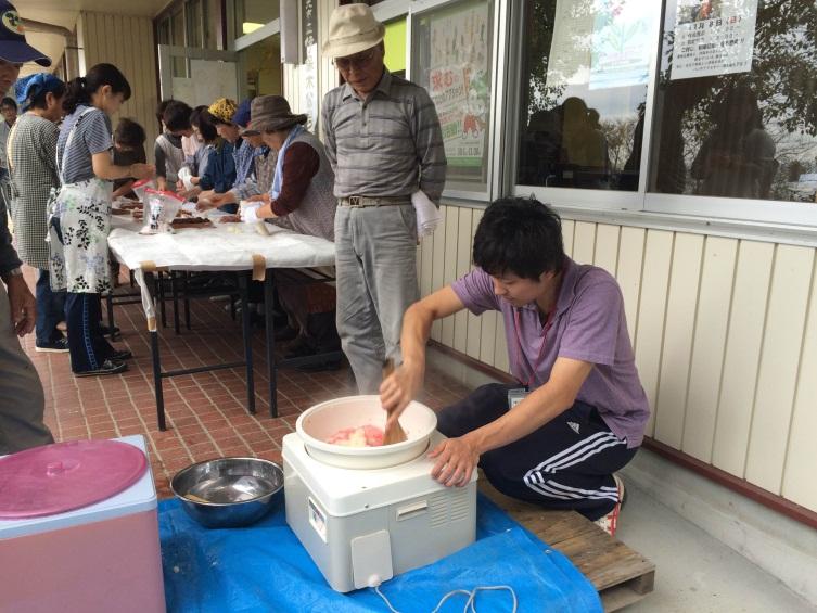 公民館で餅つき機の中味をしゃもじで混ぜている男性を帽子を被った男性が見ている。その横のテーブルでエプロン姿の女性たちが餅を丸めてる写真