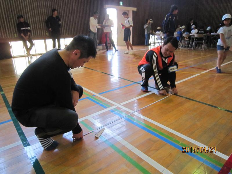 体育館で女の子が飛ばした紙飛行機の距離をメジャーで測っている男性隊員の写真