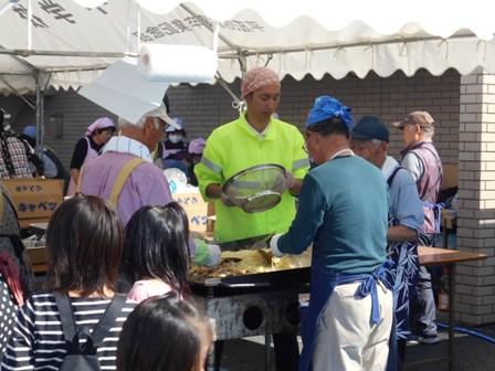 テントの中にある鉄板でスタッフの男性たちが焼きそばを作っているようすをお客が見ている写真