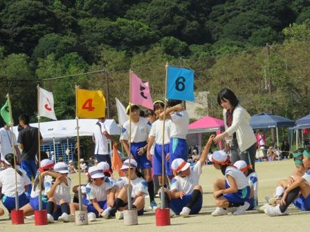 グラウンドで番号の旗が立てられている後ろに帽子やハチマキをつけている体操服姿の学生たちが座っている写真