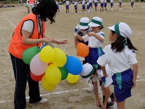 グラウンドで帽子に鉢巻きを付けた体操服姿の小学生たちに女性隊員が競技用の風船を配布している写真