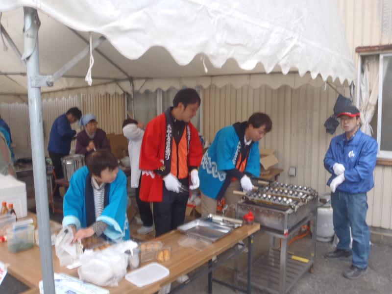 赤や、青の法被を着て、テント内で祭りの準備をする関係者の写真