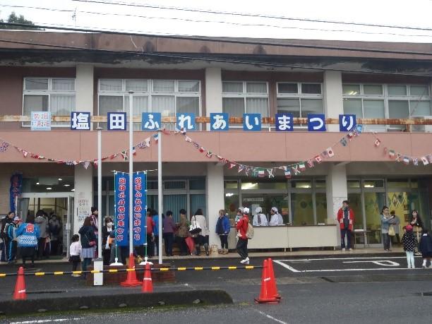 塩田コミュニティーセンターの正面に「塩田ふれあいまつり」の文字と、のぼり旗が飾られており法被姿の隊員や人々が写っている写真