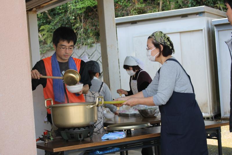 男性隊員が鍋に入った料理をおたまで器についでいるのを御盆を手に持って待っている女性スタッフの写真