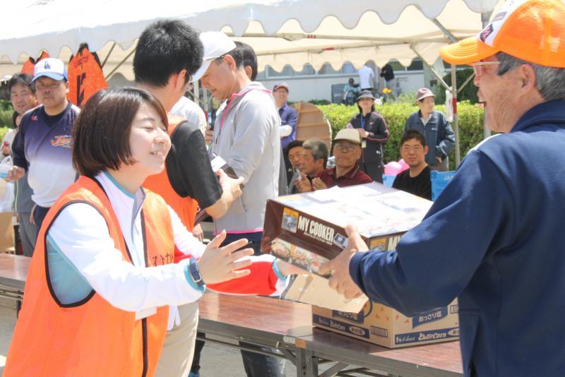 男性スタッフより景品を笑顔で受け取る女性隊員をテントの中から見ている人々の写真