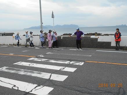 横断歩道の先の海沿いで資料を見ながら歩くウォークラリーの参加者たちを隊員の女性が見守っている写真