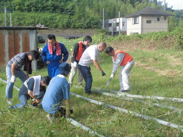 草むらに長い竹が置かれており、男性7名で相談しながら竹に細工をしている写真