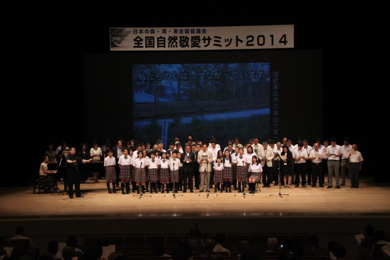 壇上で大勢の協議会会員と関係者が歌っている写真