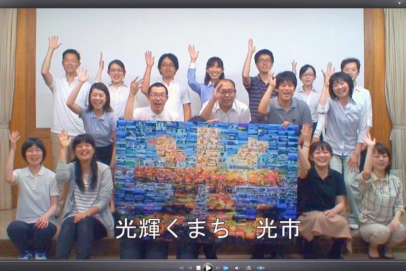 大勢の人が3列に並び右手を軽く上げているシティセールス用ビデオの1場面のキャプチャ