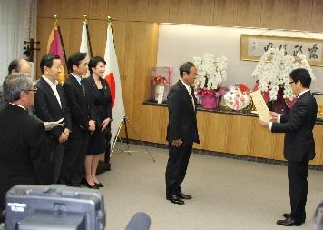 賞状のようなものを手に持った男性の前に背筋を伸ばして立つ男性の写真