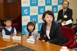 男の子と女の子、2人の子どもを連れたスーツ姿の女性が発言している写真