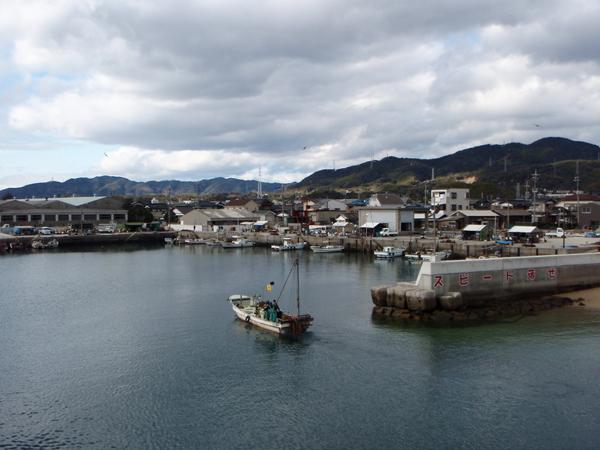 山を背景に並ぶ古い建物がたくさんの港の赤い字でスピード落とせと書かれた防波堤の間を通っている白い漁船