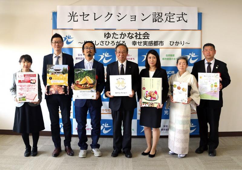 光セレクション認定式と書かれた横断幕のしたで、商品画像の印刷された色紙を手にカメラのほう向いて立つ人たちの写真