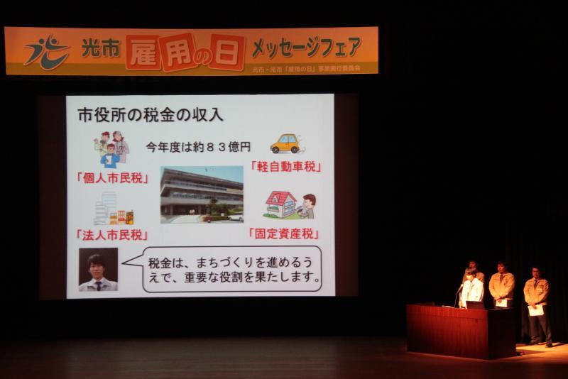ステージ上のスクリーンに市役所の税金について映し出されている写真
