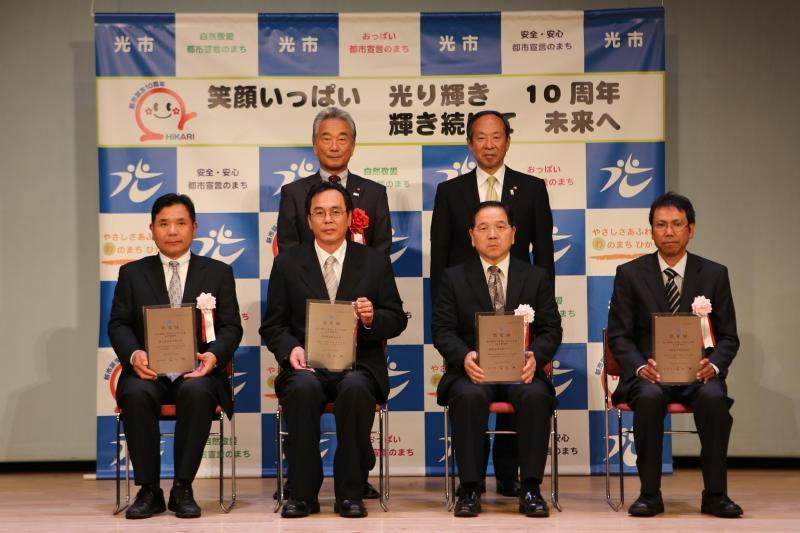 室内で手前に座って認定盾を持つ四人の男性と奥で立っている二人の男性の記念写真