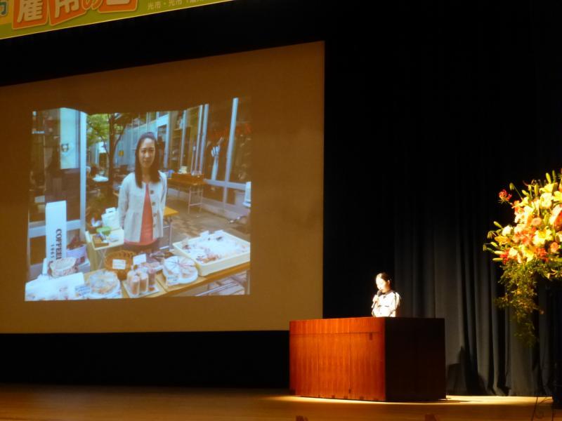ステージ上のスクリーンに屋外でお菓子を売る女性が映し出されている写真