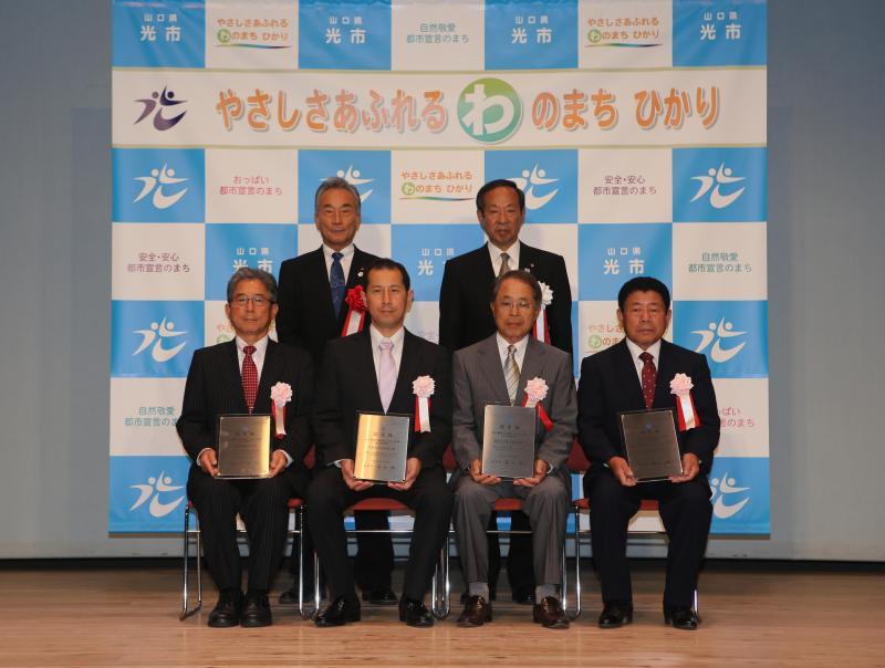 室内で手前に座って認定盾を持つ四人の男性と奥で立っている二人の男性の集合写真