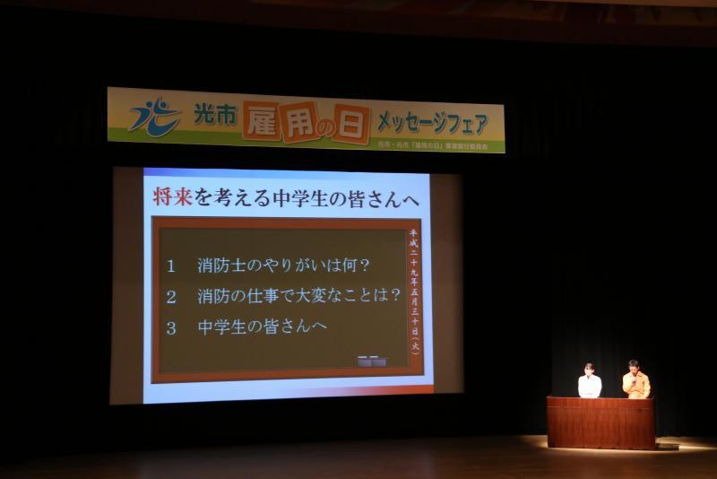 ステージ上のスクリーンに「将来を考える中学生の皆さんへ」と題されたスライドが映し出されている写真