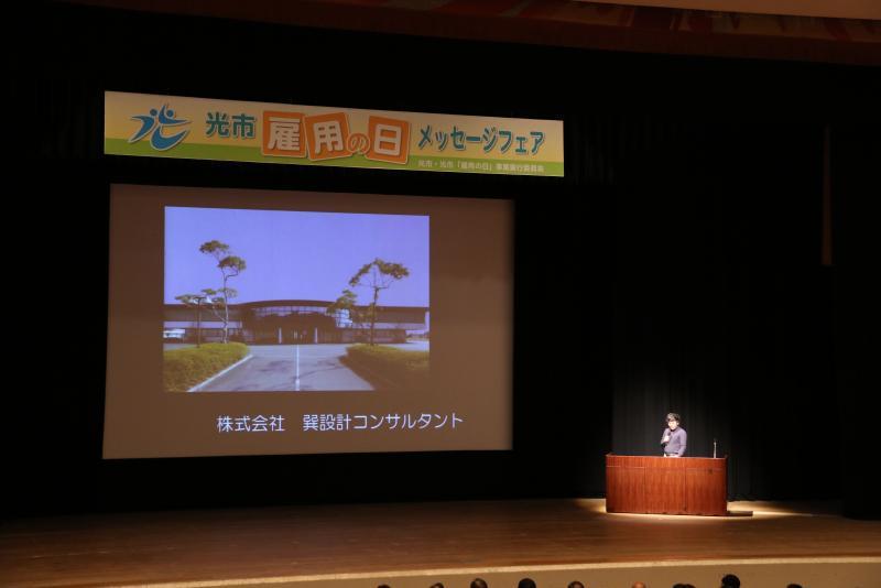 ステージ上のスクリーンに巽設計コンサルタントの建物外観が映し出されている写真